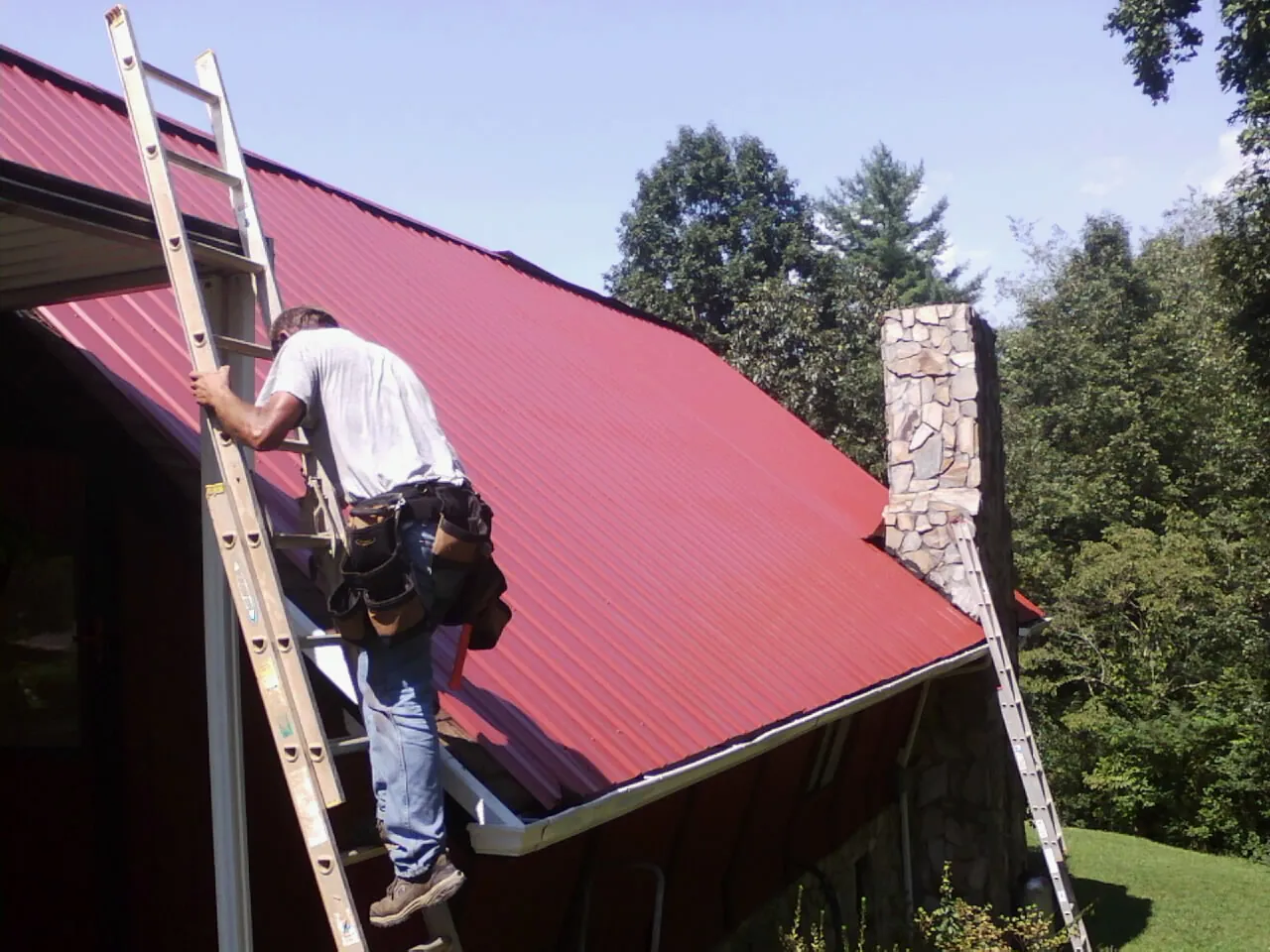 Roofer on a ladder beside a red metal roof and stone chimney — Soffit & Fascia Repair by Roofing Brothers in Moxee